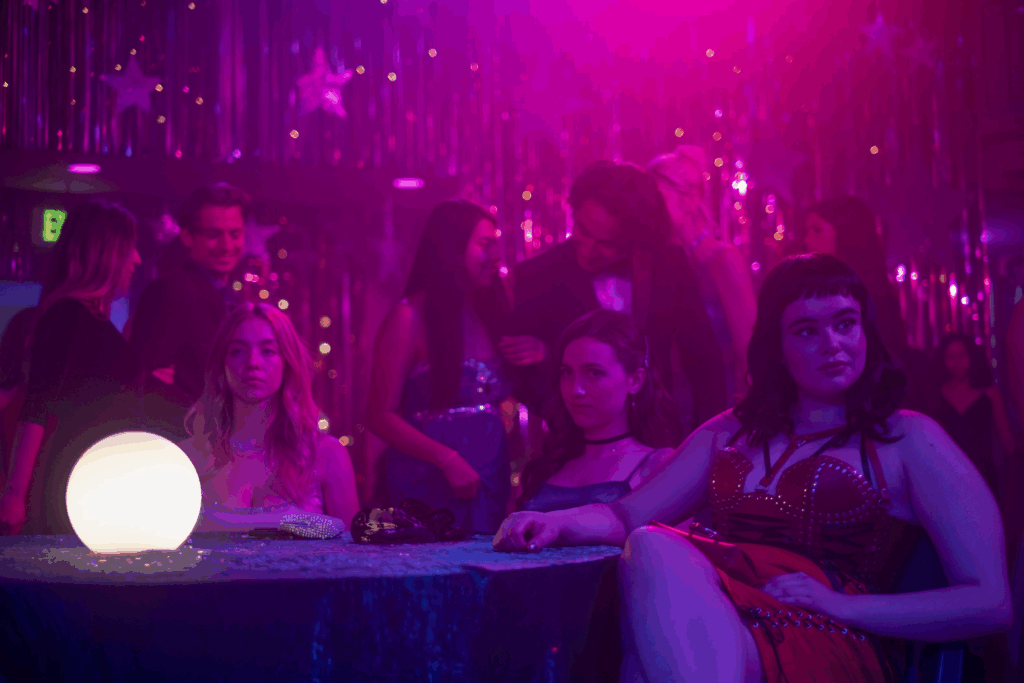 Cassie, Kat, and other students sit and mingle at a school dance decorated with purple lights and silver streamers in a scene from Euphoria