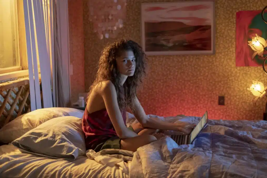 Rue Bennett (portrayed by Zendaya) sitting on a bed using a laptop in a dimly lit bedroom scene from Euphoria
